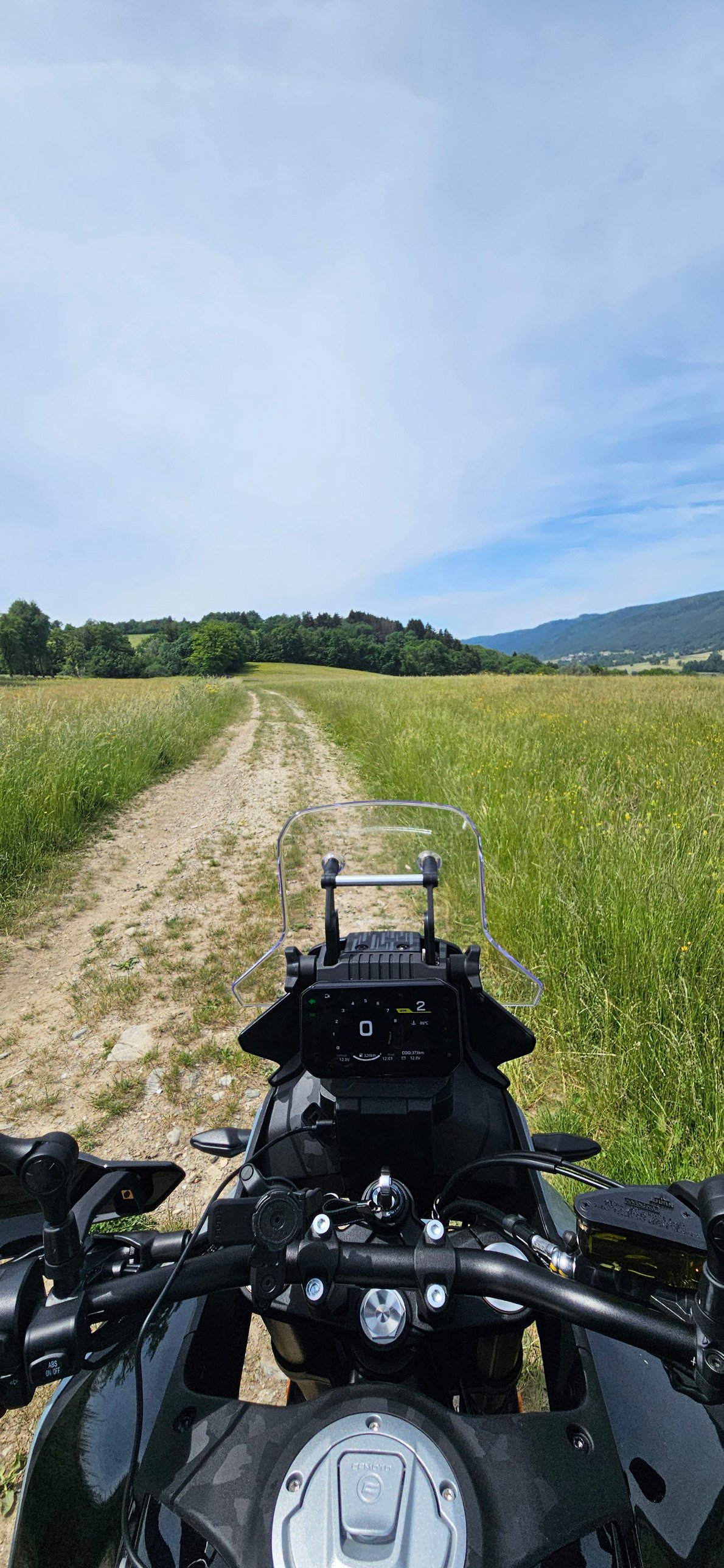 Vue depuis le guidon d'une CFMOTO 450MT sur un chemin de terre entre deux prés, forêt et montagnes à l'horizon